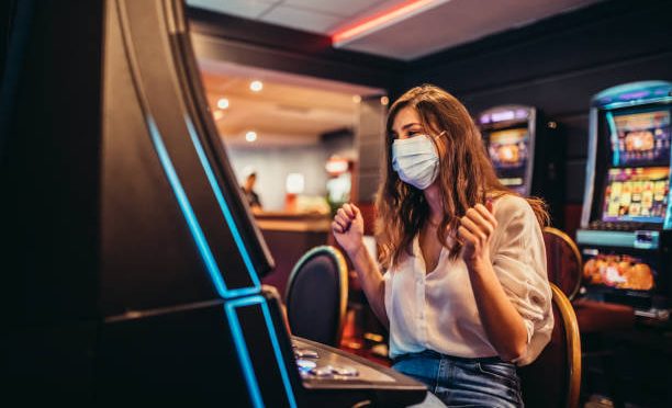 Beautiful young woman with protective face mask playing slot machine in casino.